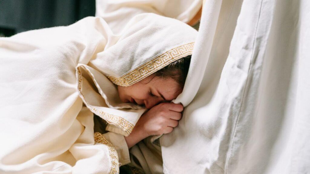 Mulher ajoelhada em oração, envolta em vestes claras, expressando fé, dor e esperança